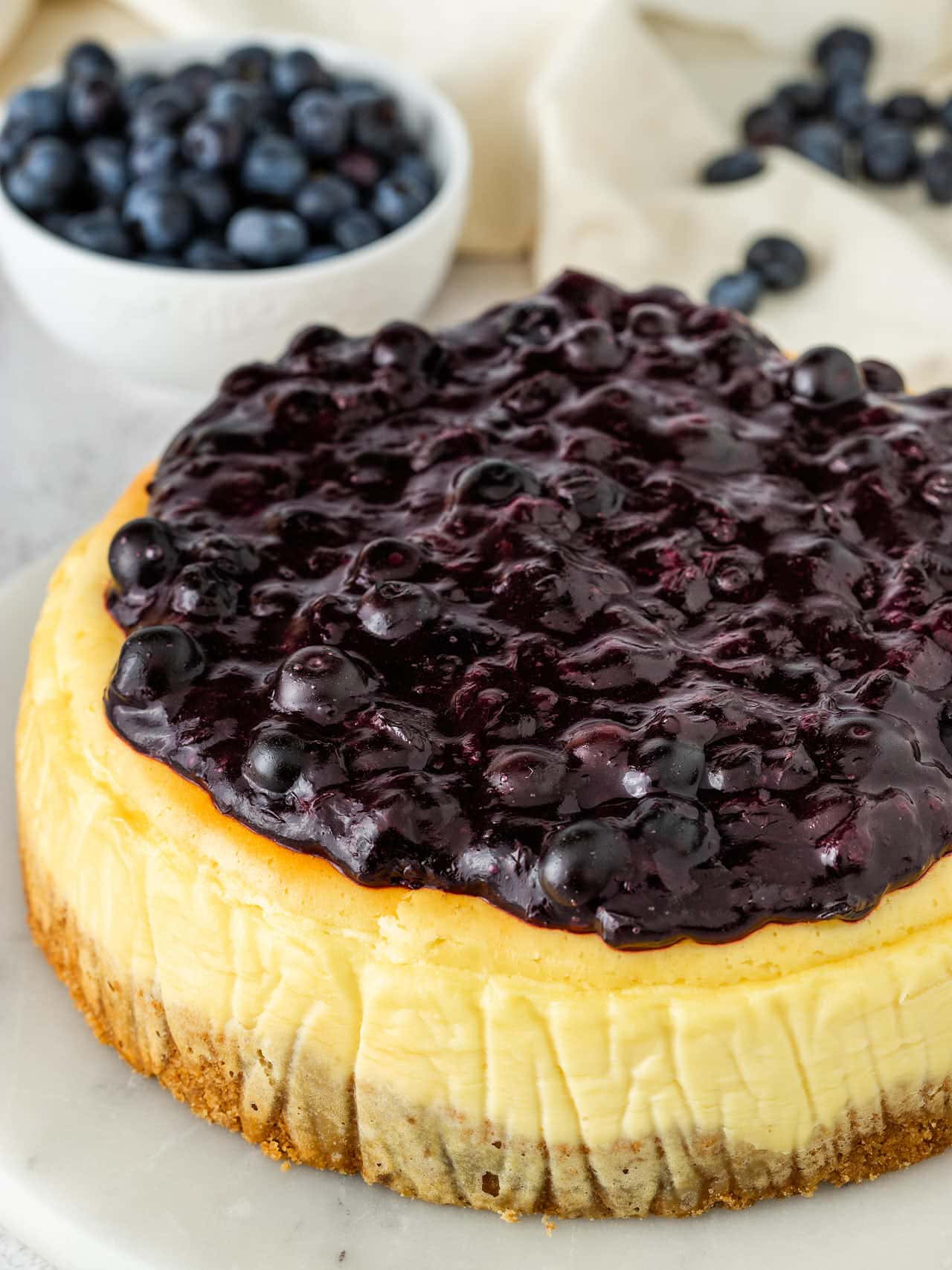 A whole Blueberry Cheesecake topped with thick blueberry sauce, with a bowl of fresh blueberries behind it.