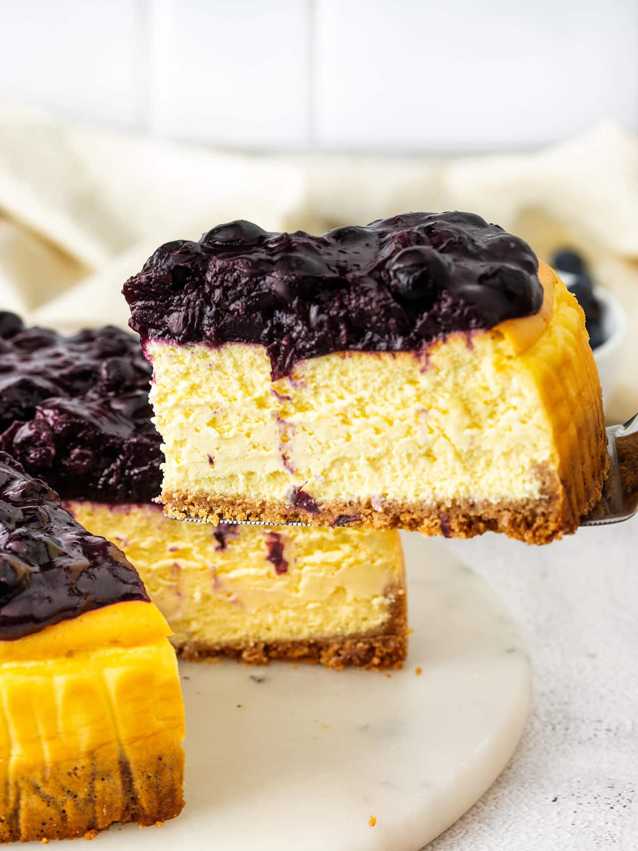 A slice of Blueberry Cheesecake lifted on a server, showing the creamy filling, graham cracker crust, and berry topping.