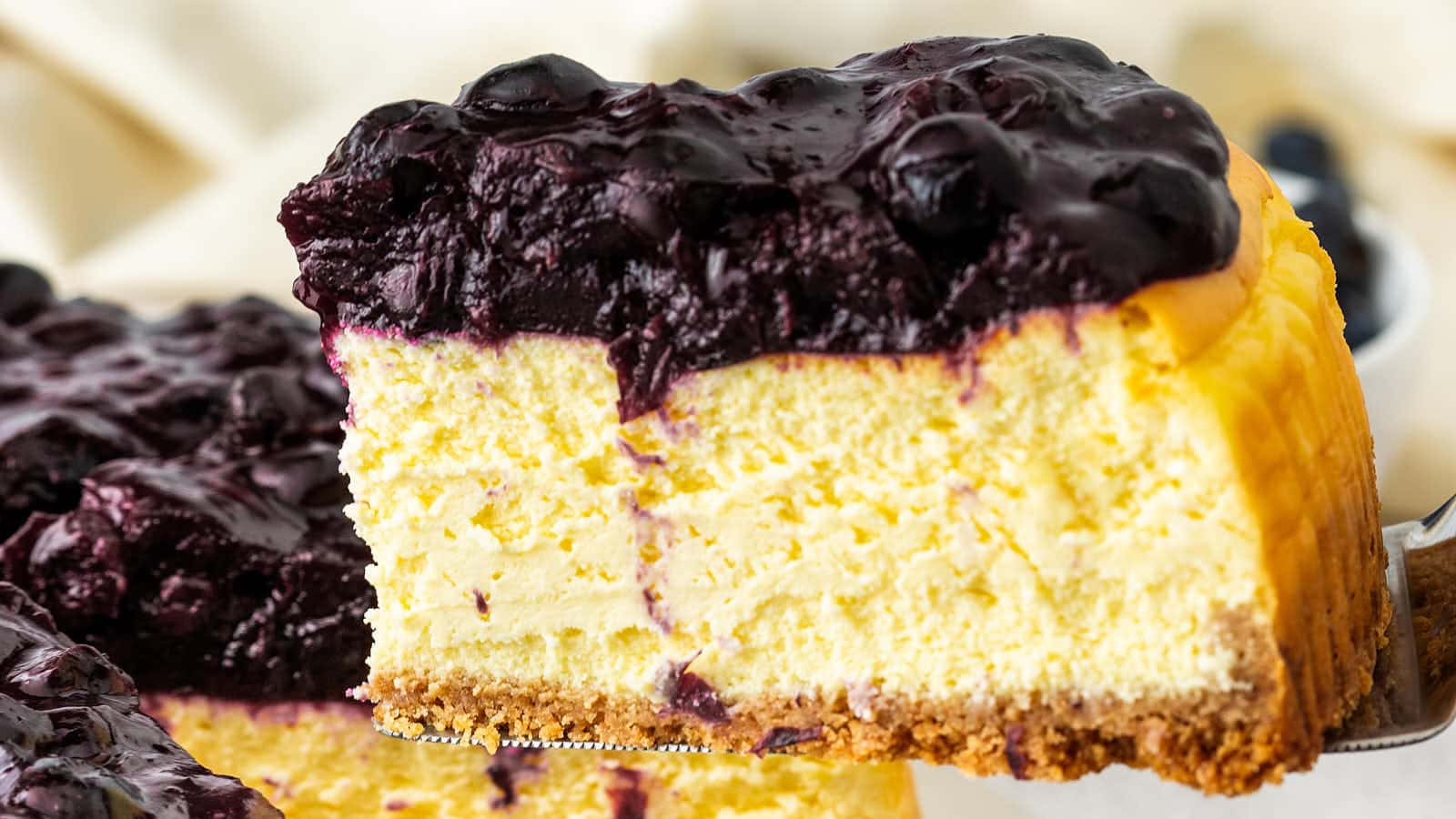 A slice of Blueberry Cheesecake lifted on a server, showing the creamy filling, graham cracker crust, and berry topping.