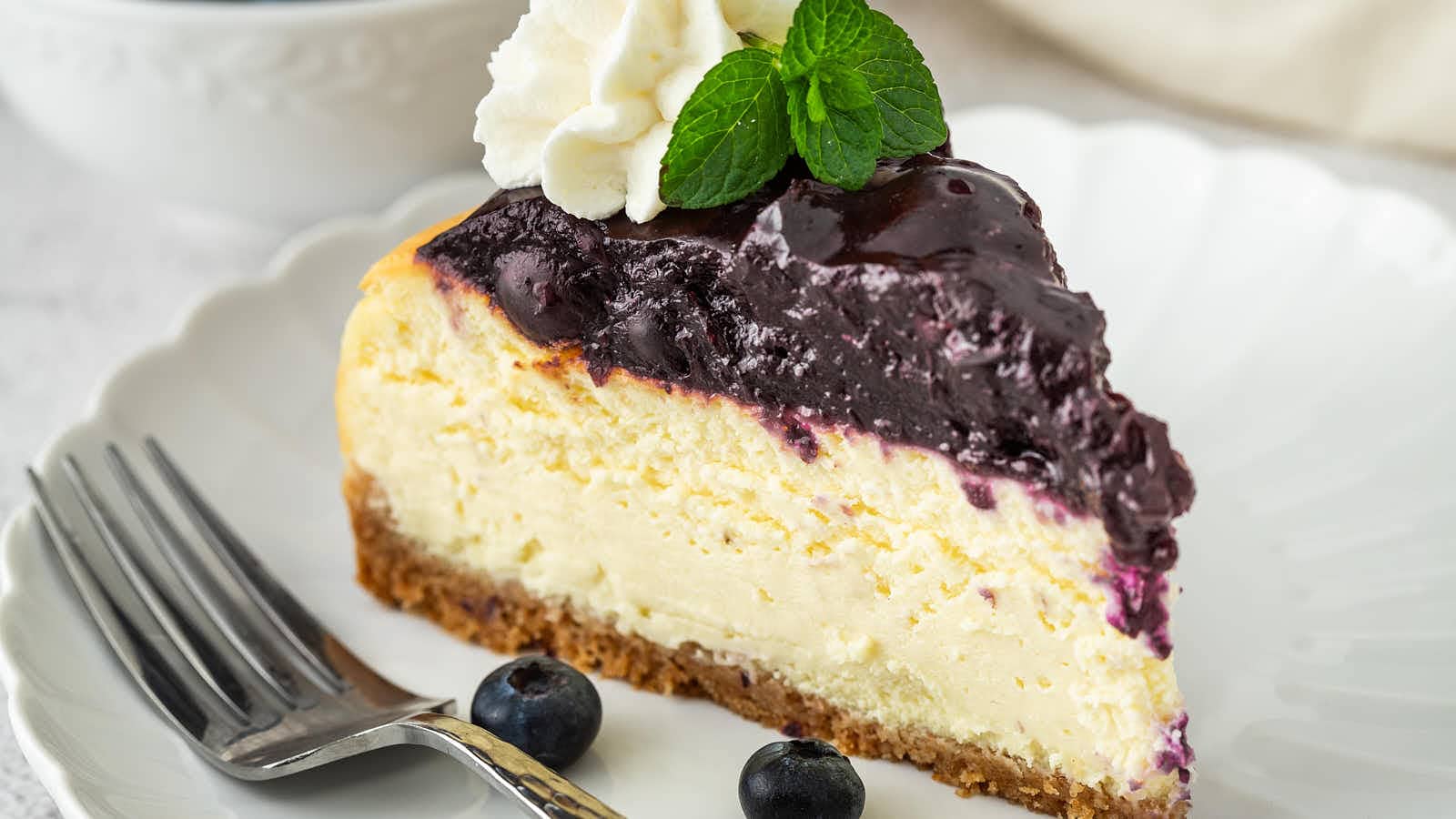 A slice of Blueberry Cheesecake topped with whipped cream and a mint sprig, with a fork and fresh blueberries.