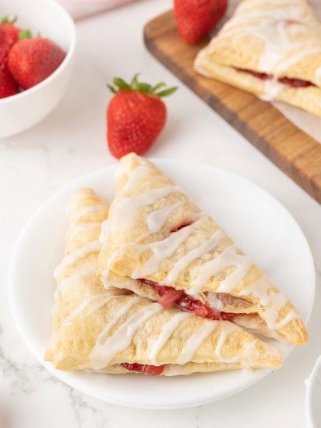 Two glazed Strawberry Turnovers on a white plate with fresh strawberries.