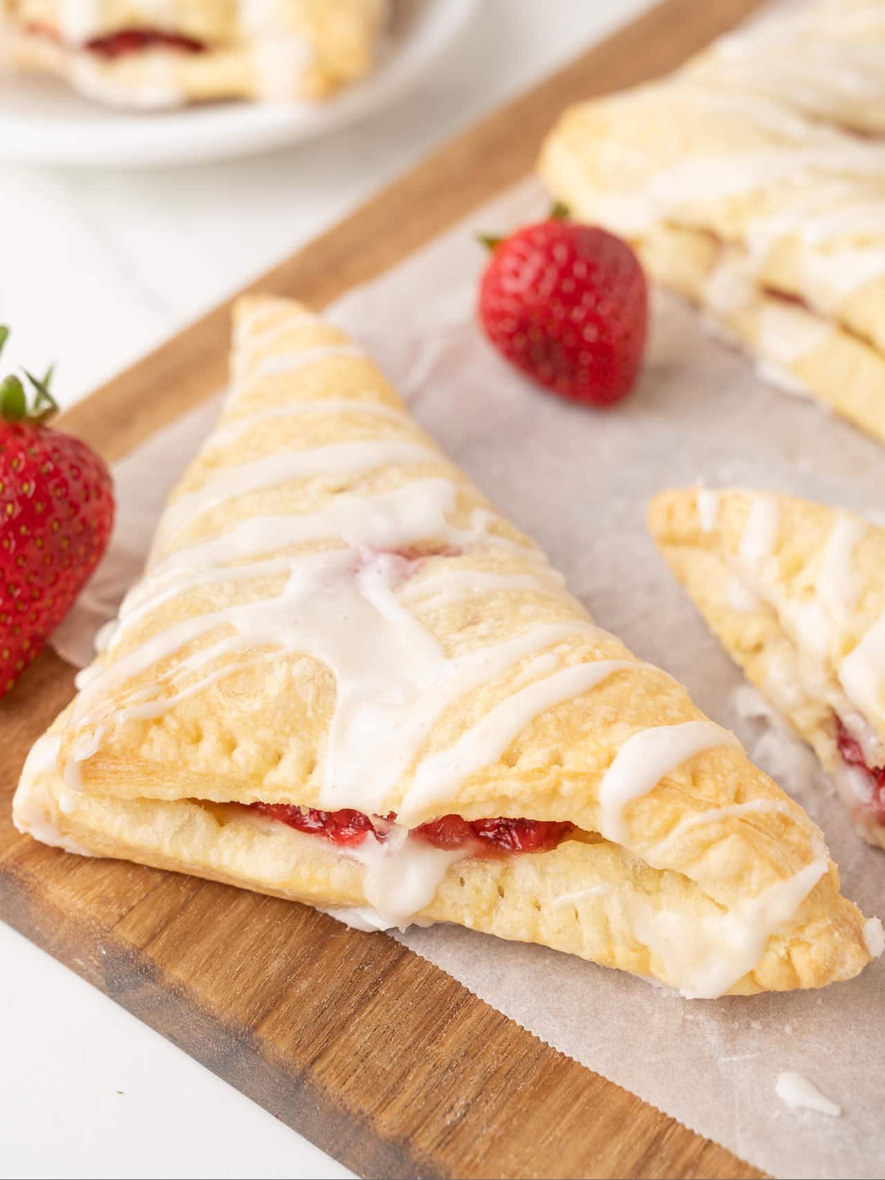 A Strawberry Turnover with an icing drizzle on a wood board, with fresh strawberries beside it.