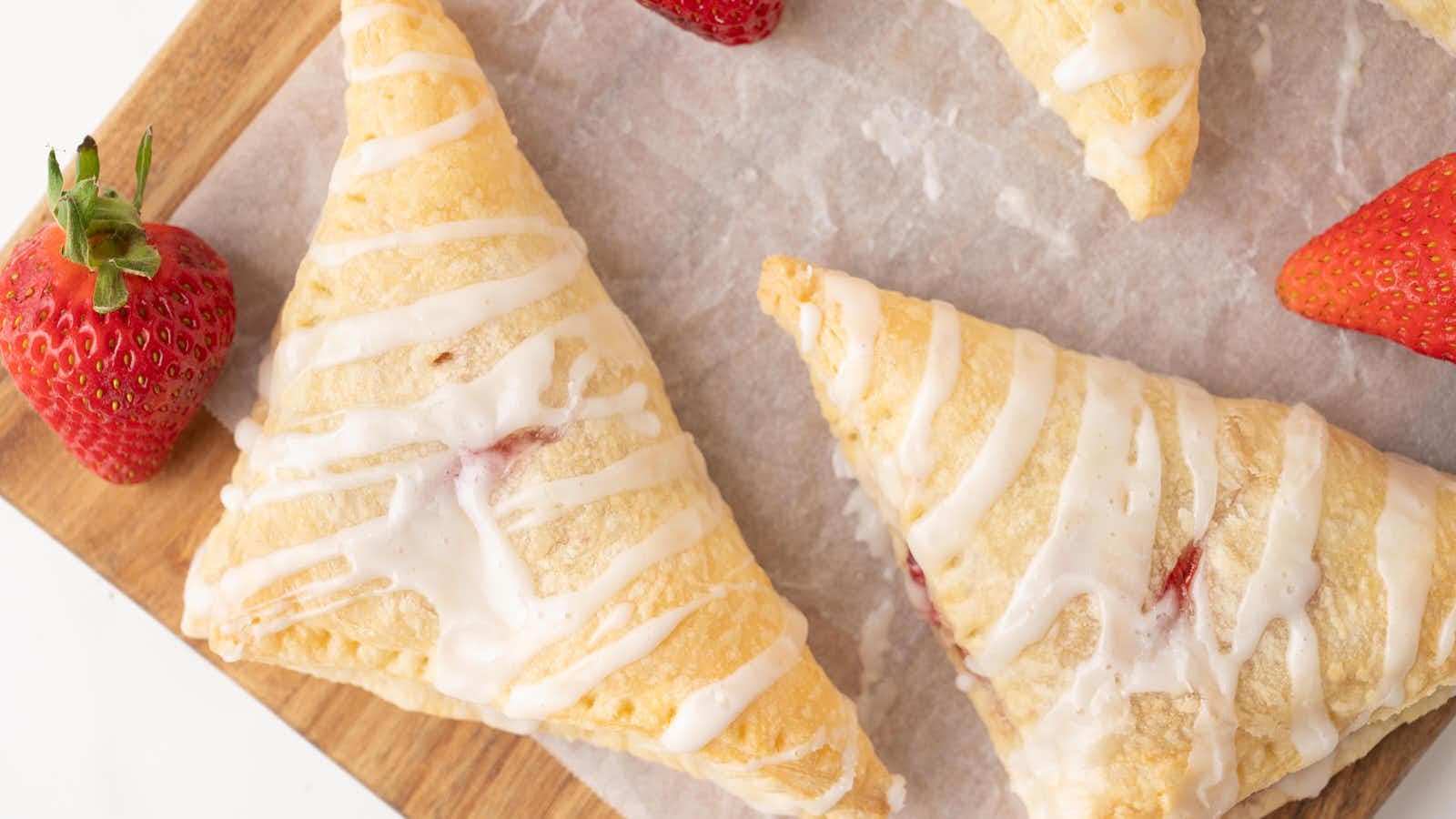 Overhead close-up of glazed Strawberry Turnovers on a wood board with a fresh strawberry.