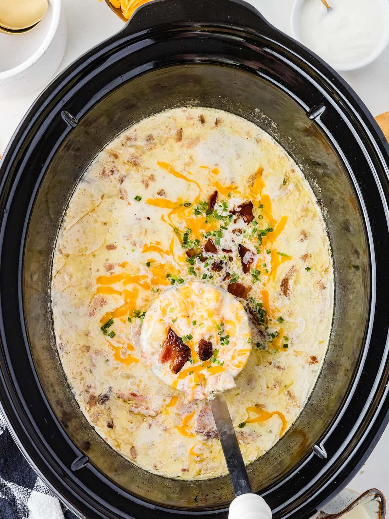 Ladle lifting creamy Crockpot Loaded Potato Soup from a black slow cooker.