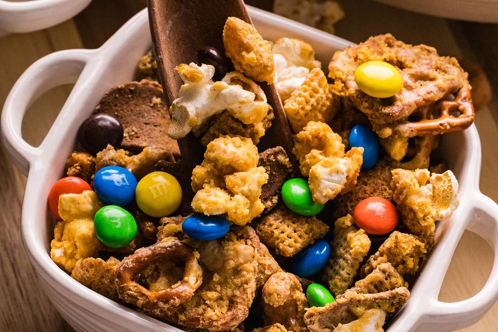 Square dish of Sweet and Salty Chex Mix with a wooden spoon, surrounded by small bowls.