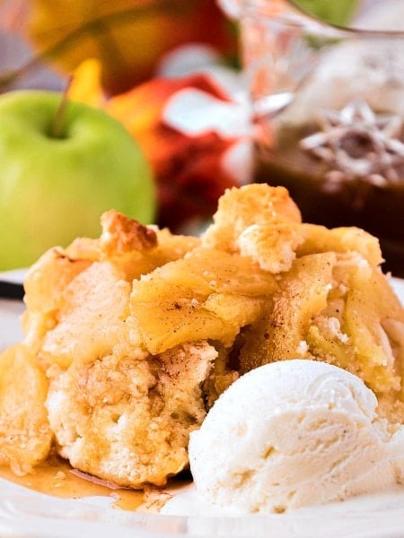 A serving of Apple Cobbler with vanilla ice cream, apples, and caramel sauce in the background.