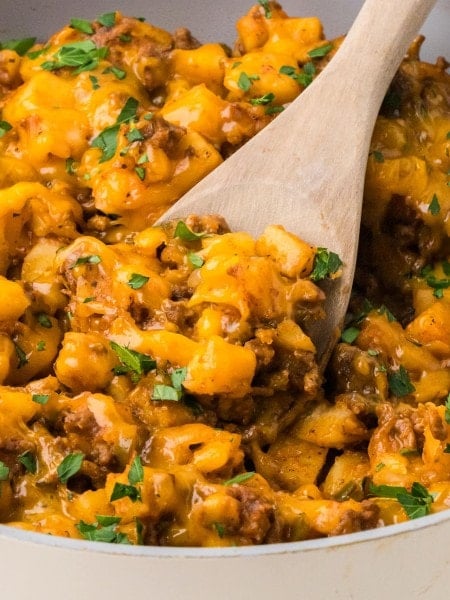Wooden spoon scooping cheesy Ground Beef and Potatoes from a skillet.