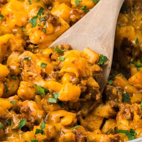 Wooden spoon scooping cheesy Ground Beef and Potatoes from a skillet.