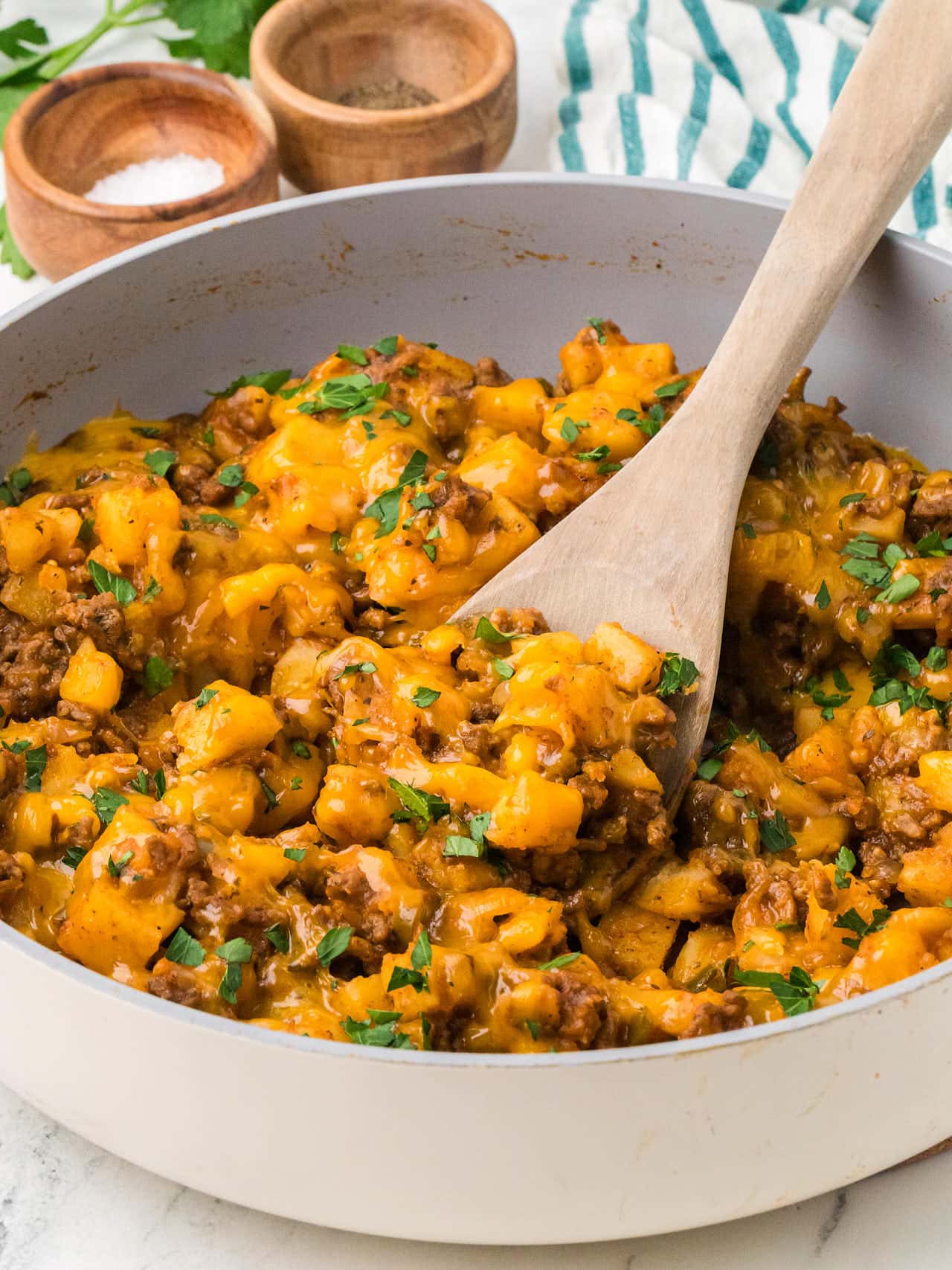 Side view of a skillet of Ground Beef and Potatoes with melted cheddar and a wooden spoon.