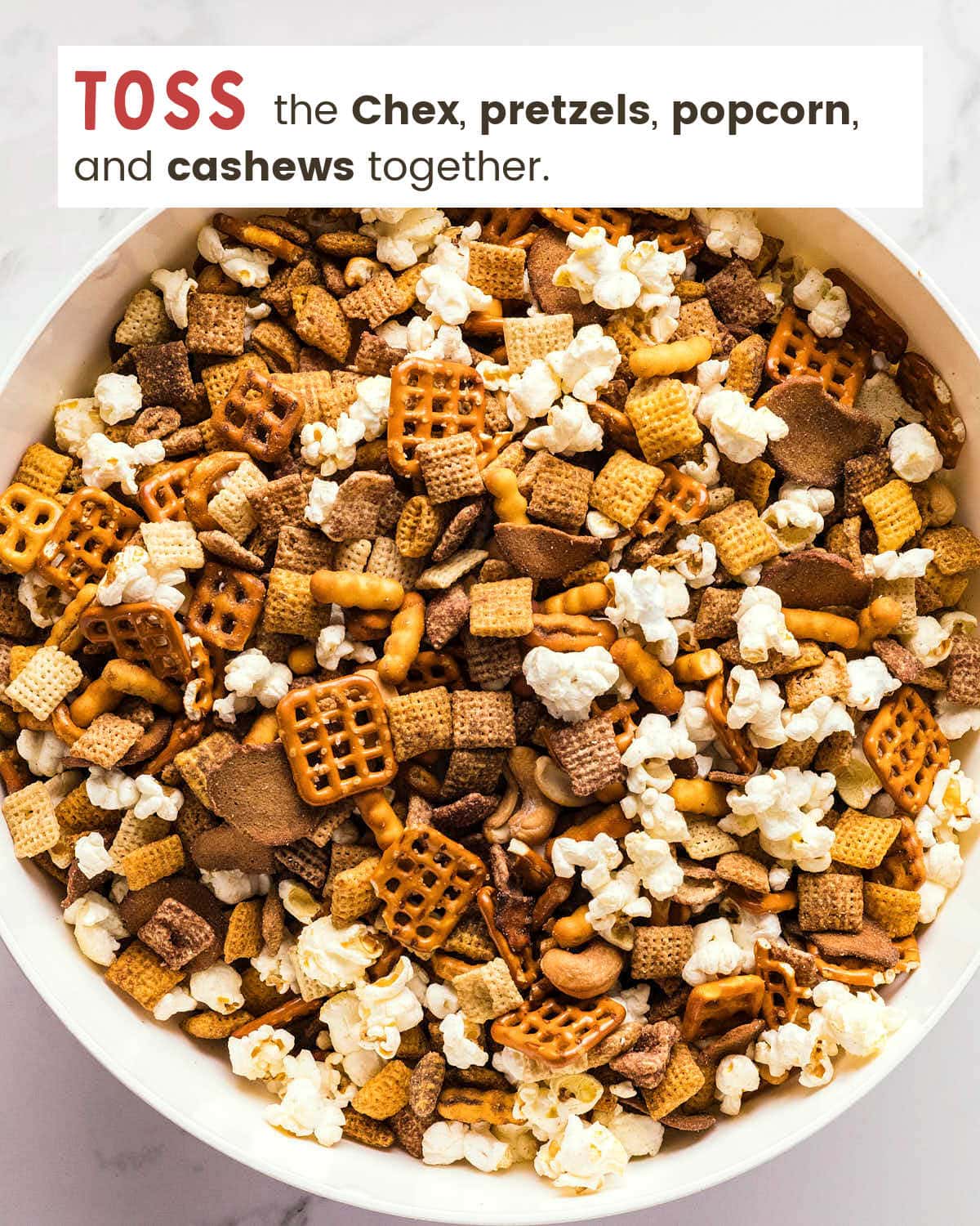 Chex cereal, pretzels, popcorn, and cashews tossed together in a white bowl.