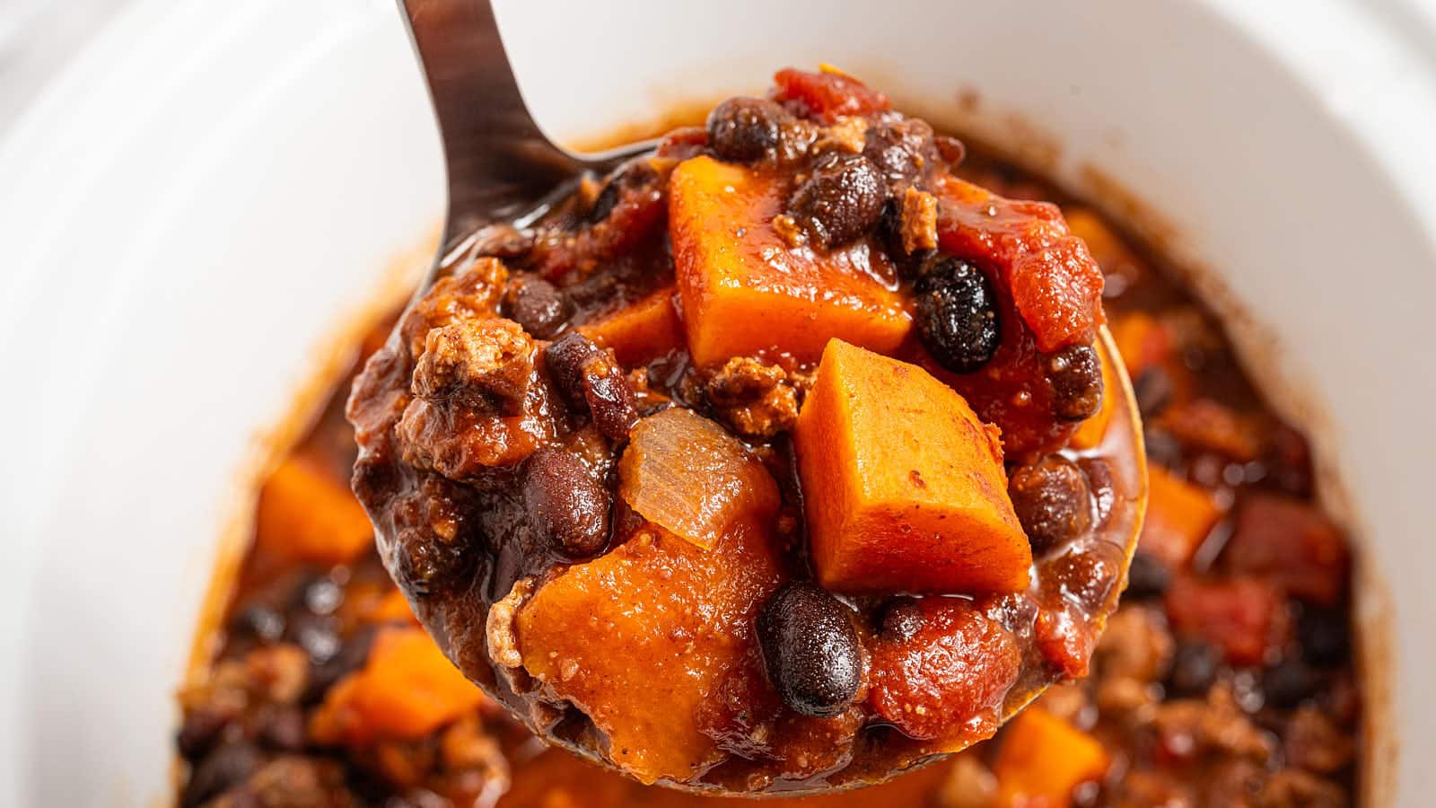 Ladle of Crockpot Ground Turkey Chili with sweet potato cubes, black beans and tomatoes over the slow cooker.