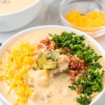 A bowl of Cheeseburger Soup topped with shredded Cheddar, bacon bits, diced pickles, green onions, and parsley.