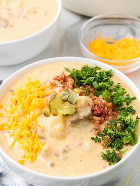 A bowl of Cheeseburger Soup topped with shredded Cheddar, bacon bits, diced pickles, green onions, and parsley.