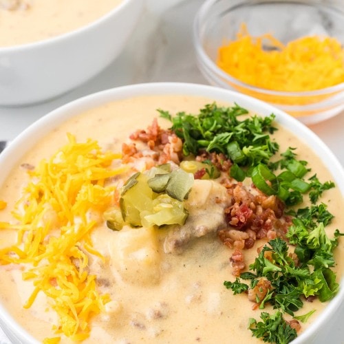 A bowl of Cheeseburger Soup topped with shredded Cheddar, bacon bits, diced pickles, green onions, and parsley.