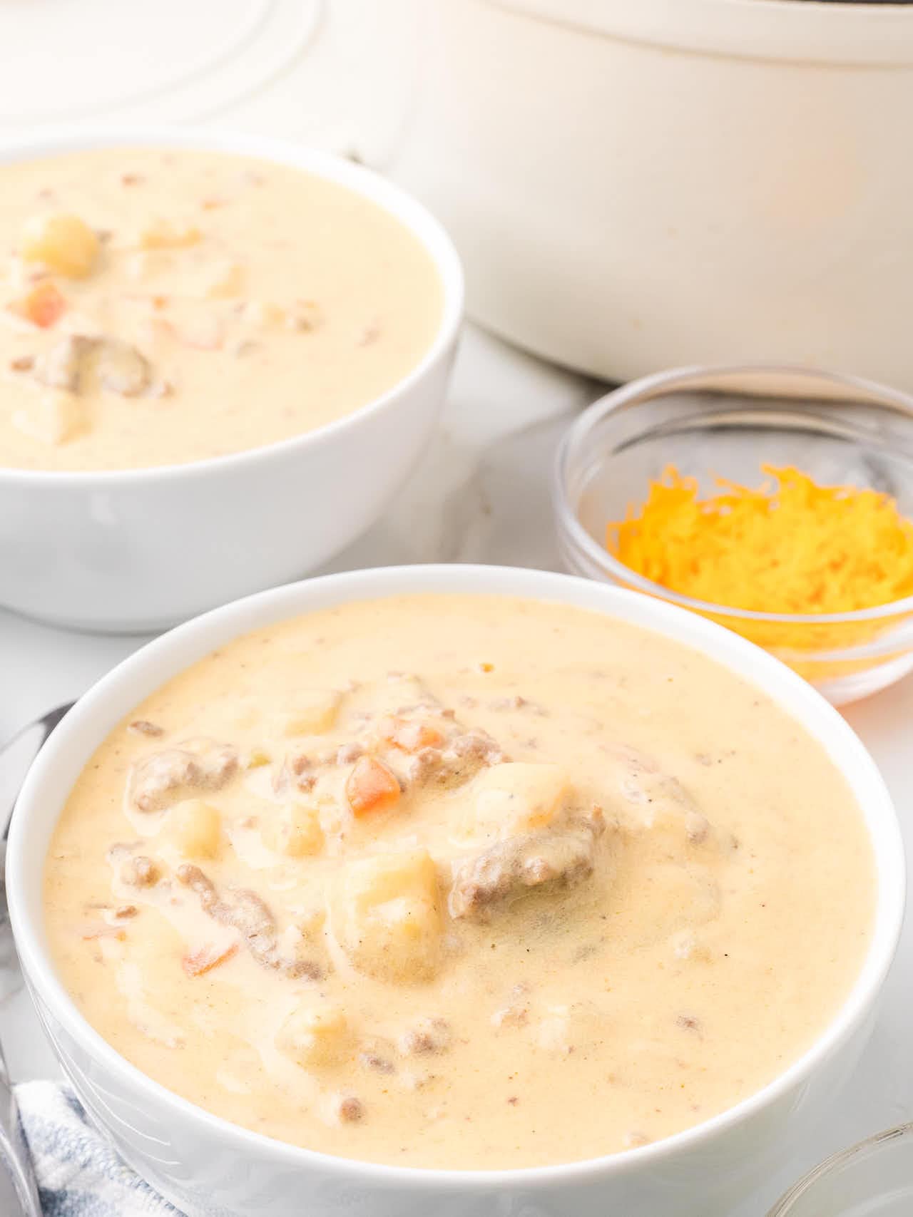 Two white bowls of Cheeseburger Soup next to a small dish of shredded Cheddar.