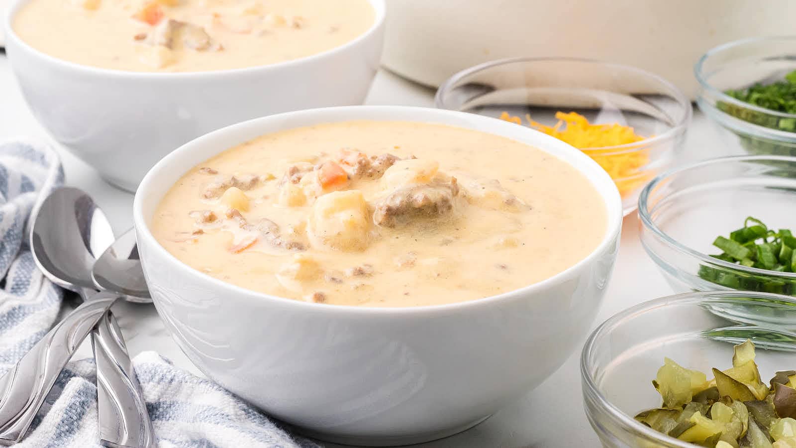 Side view of two bowls of Cheeseburger Soup with spoons and toppings in the background.