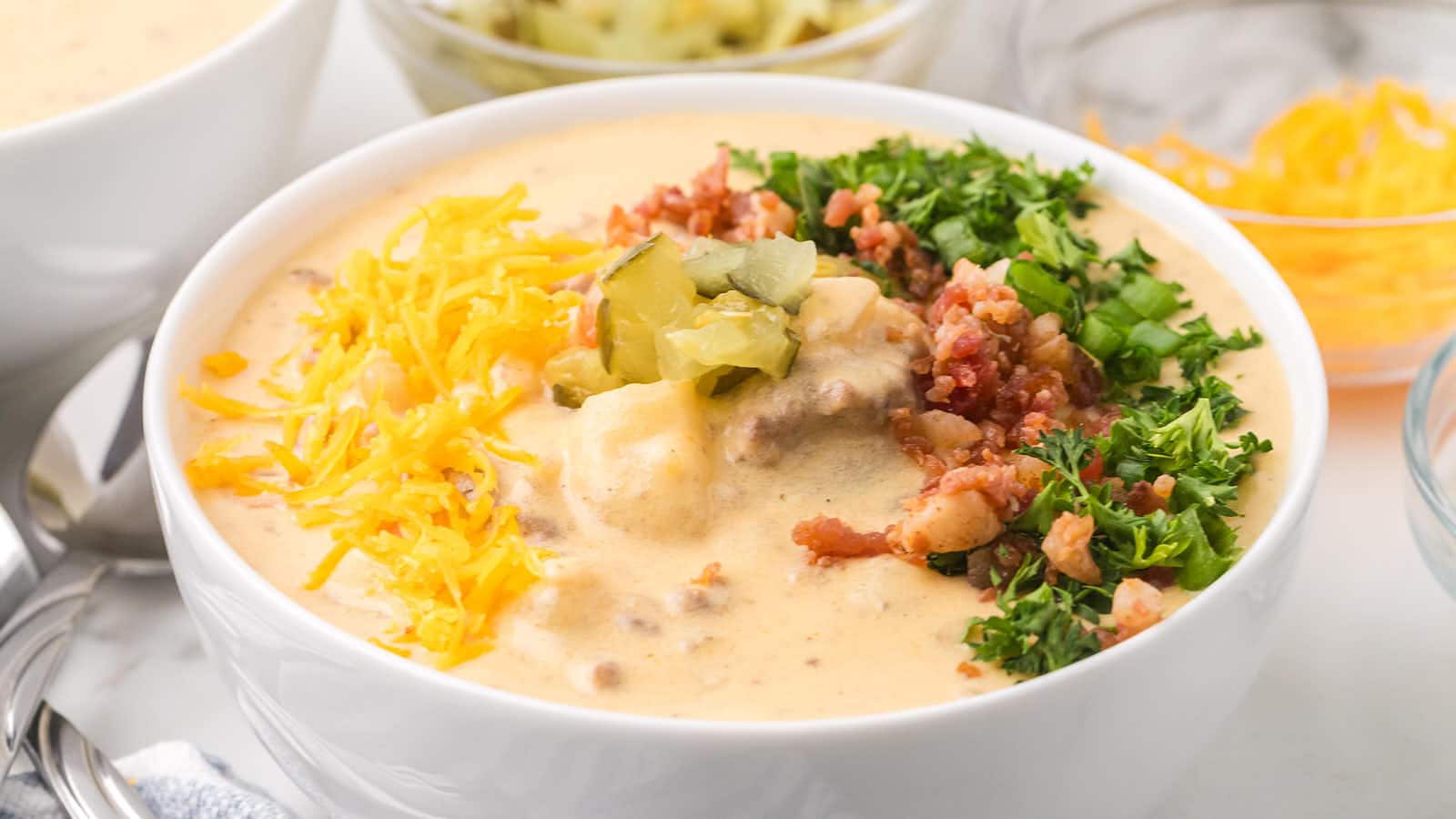 A loaded bowl of Cheeseburger Soup with Cheddar, bacon, pickles, and parsley on top.