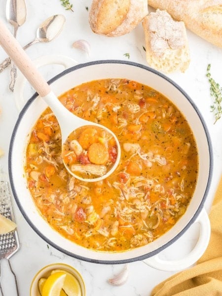 A ladle lifting Chicken and White Bean Soup from a white Dutch oven, with crusty bread and lemon wedges nearby.