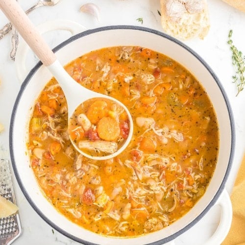 A ladle lifting Chicken and White Bean Soup from a white Dutch oven, with crusty bread and lemon wedges nearby.