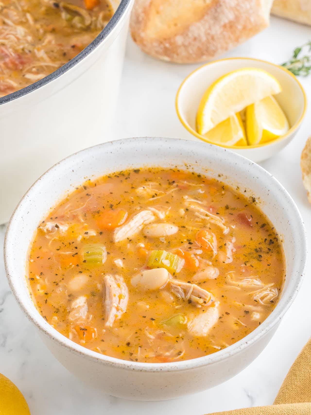 A bowl of Chicken and White Bean Soup with shredded chicken, beans, carrots and celery, with lemon wedges behind it.