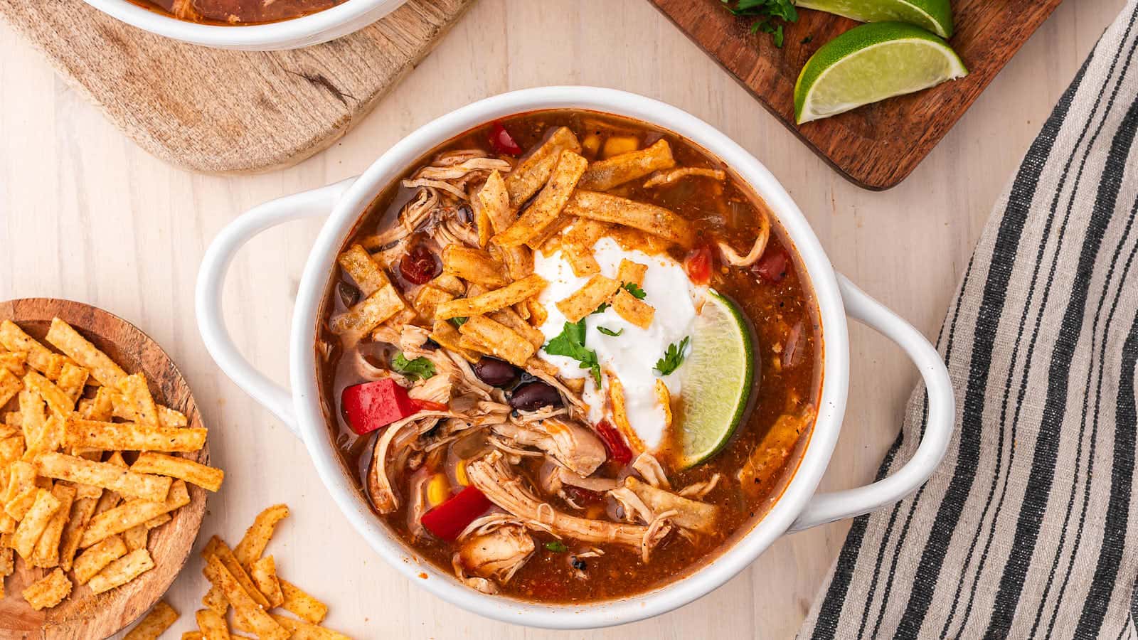Overhead view of Crockpot Chicken Enchilada Soup in a white bowl on a wooden board with fresh cilantro, lime wedges and tortilla strips.