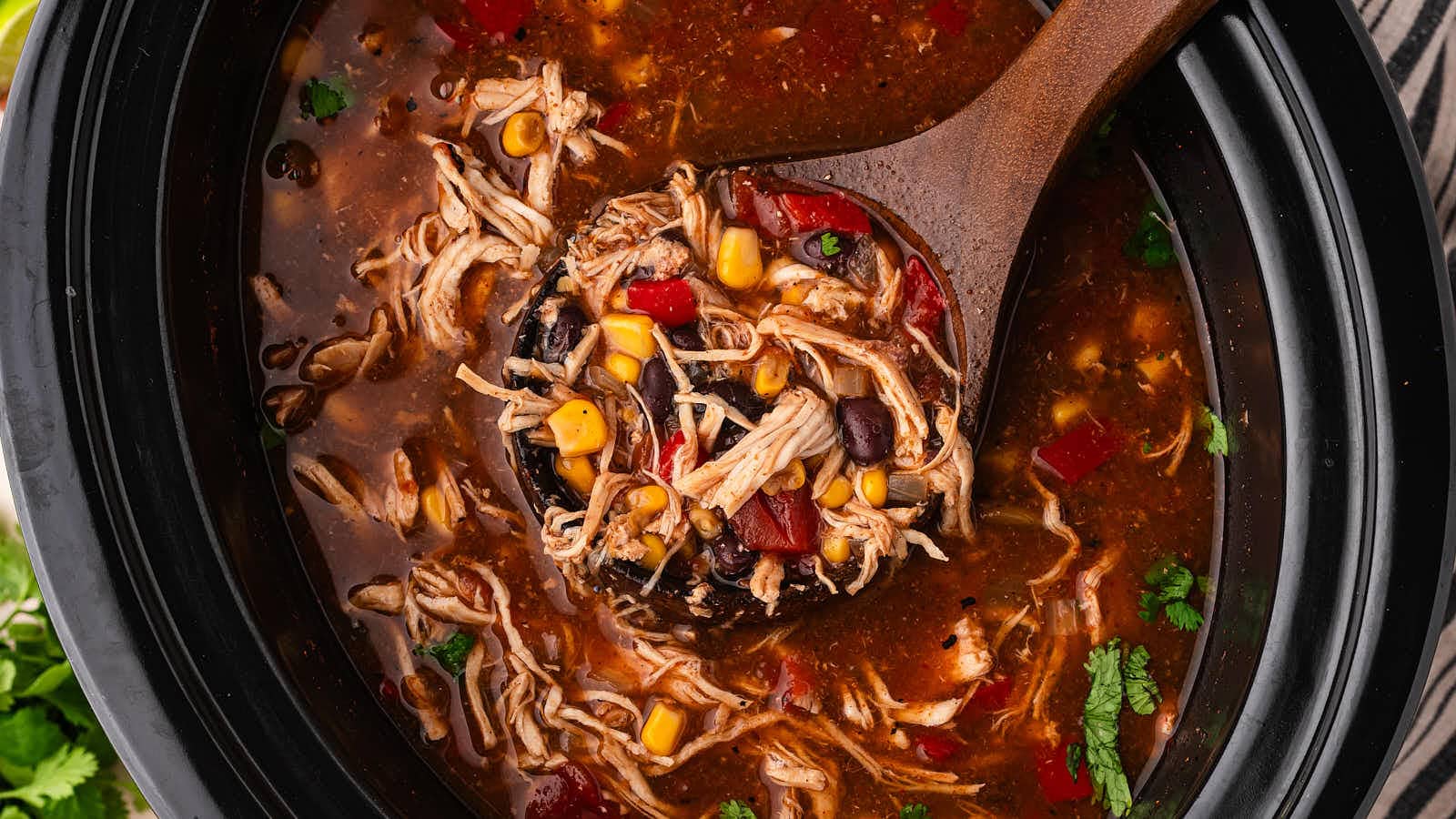 Closeup of a wooden ladle lifting shredded chicken, corn, black beans and bell pepper from Crockpot Chicken Enchilada Soup.